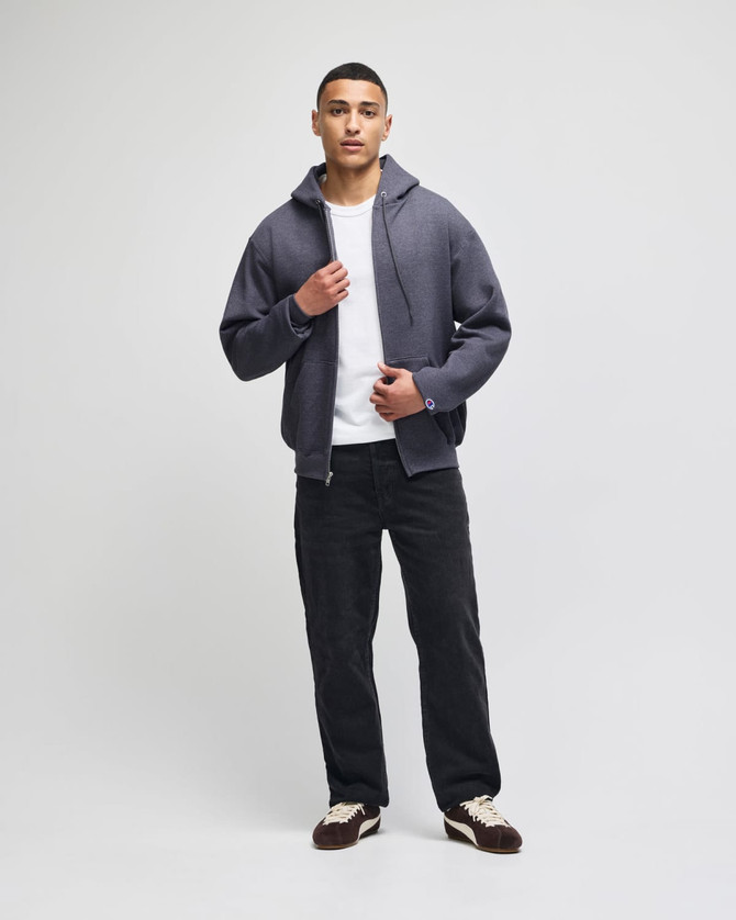 A grey zip-up sweatshirt worn over a white shirt, paired with black pants and shoes. The model stands in a neutral setting.