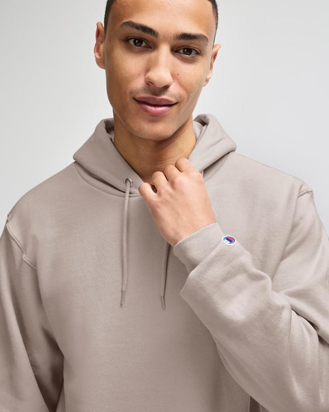 A man models a taupe Champion Powerblend® adult pullover hooded sweatshirt with a logo on the sleeve.