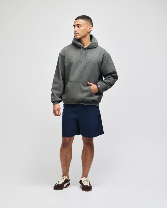 An adult pullover hooded sweatshirt in green, paired with navy shorts. The model stands confidently with one hand in the pocket.