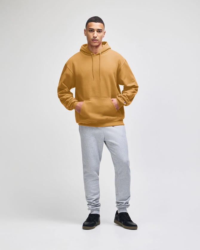 A hooded sweatshirt in a warm mustard colour paired with light grey sweatpants. The model is standing confidently.
