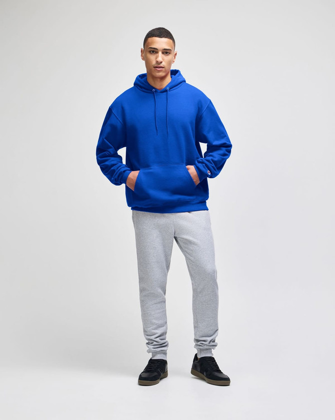 A blue pullover hooded sweatshirt with a front pocket, paired with grey sweatpants. The model stands confidently.
