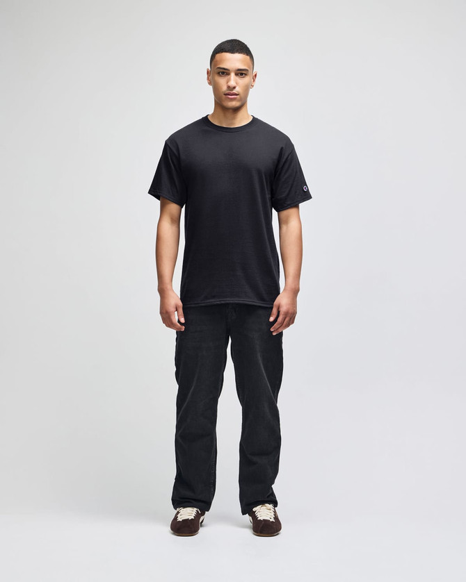 A black Champion adult short-sleeve t-shirt displayed on a model, paired with black pants. The shirt features a logo.