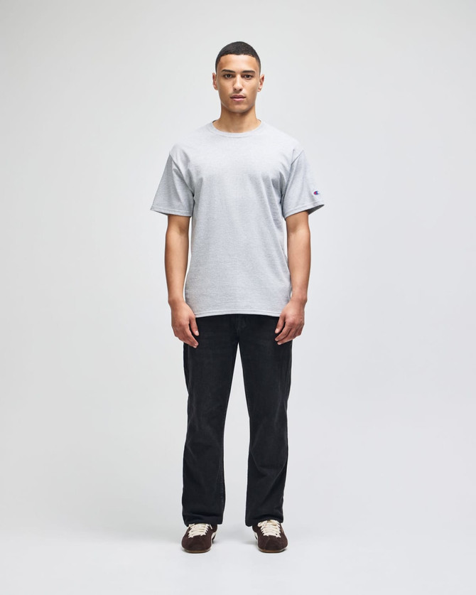 A grey short-sleeve t-shirt worn by a model, paired with dark pants. The shirt has a small logo on the sleeve.