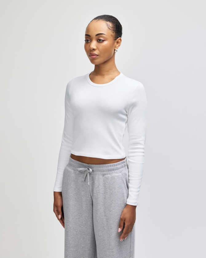 A white long-sleeve crop fitted T-shirt, modelled by a woman, paired with grey sweatpants. The shirt features a logo.