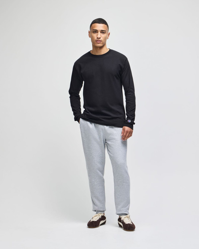 A black long-sleeve t-shirt worn with grey pants, featuring a logo on the sleeve. Model standing against a plain background.