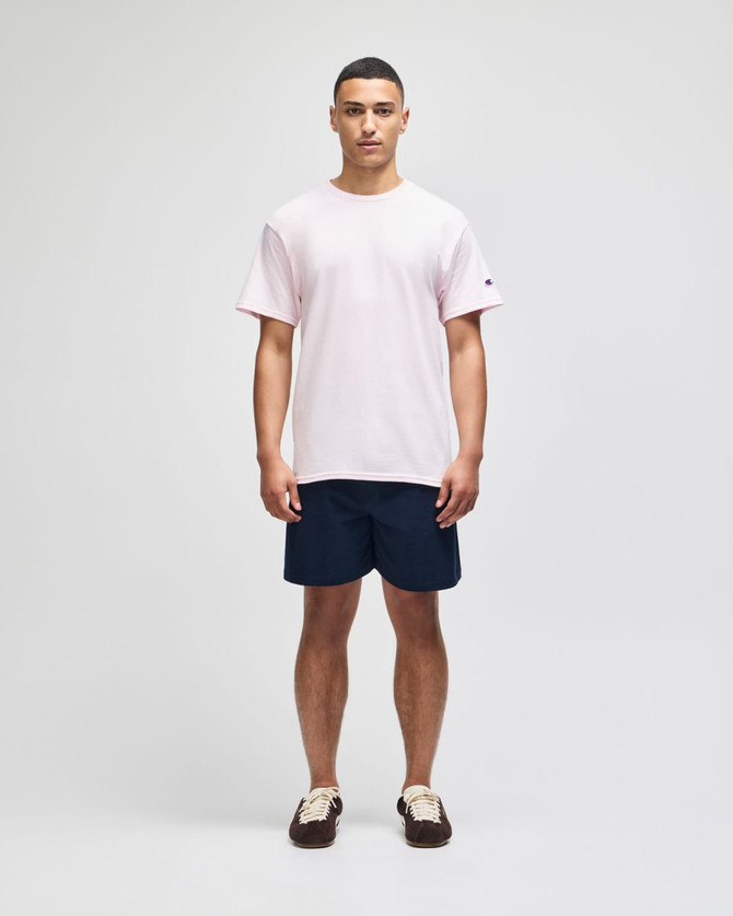 A man stands wearing a light pink short sleeve t-shirt and navy shorts, featuring a logo on the t-shirt.