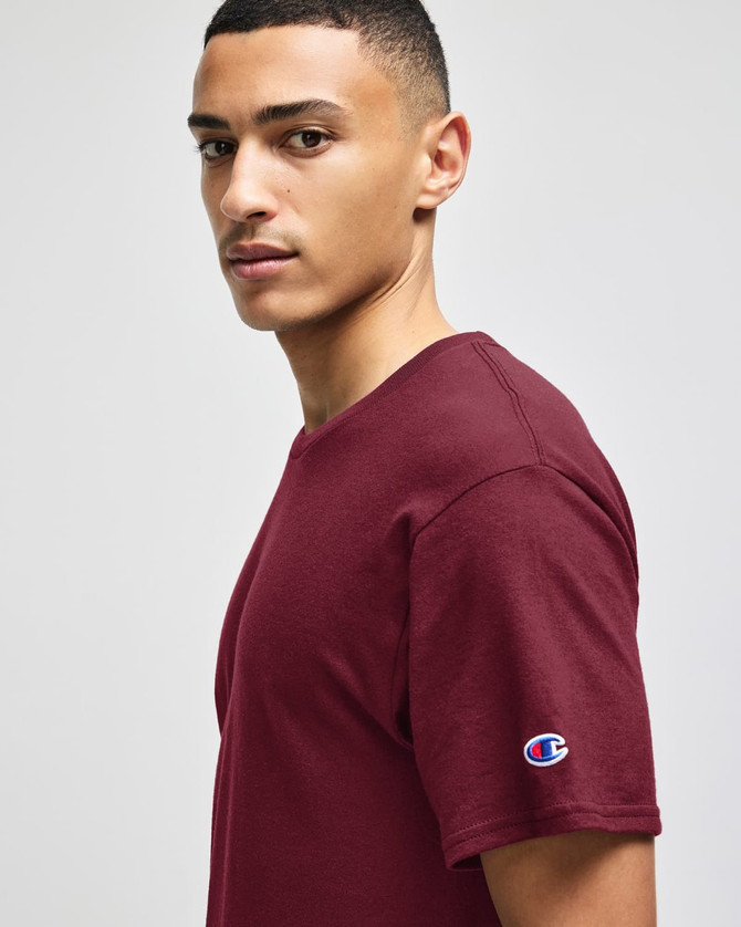 A maroon adult jersey short sleeve t-shirt with a logo on the sleeve, worn by a male model.