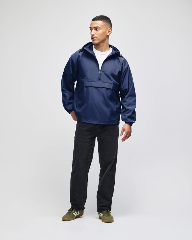 A model wearing a navy blue packable anorak jacket, paired with black trousers and green shoes. The jacket has a logo.