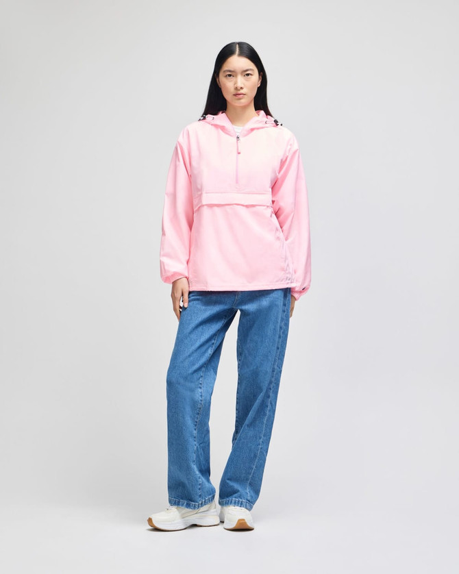 A pink packable anorak jacket with a hood, worn by a model paired with blue jeans. The jacket features a large pocket and has a logo.