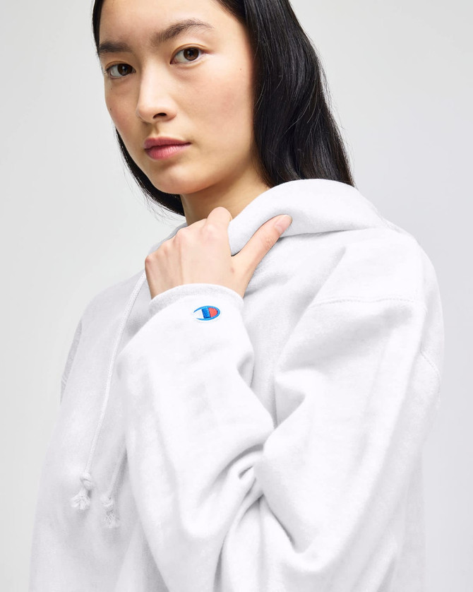 A woman wearing a white cropped hoodie with a logo on the sleeve. The hoodie features a drawstring and a relaxed fit.