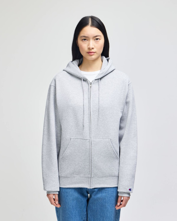 A grey full-zip hooded sweatshirt with a hood and a logo on the sleeve, styled on a model.