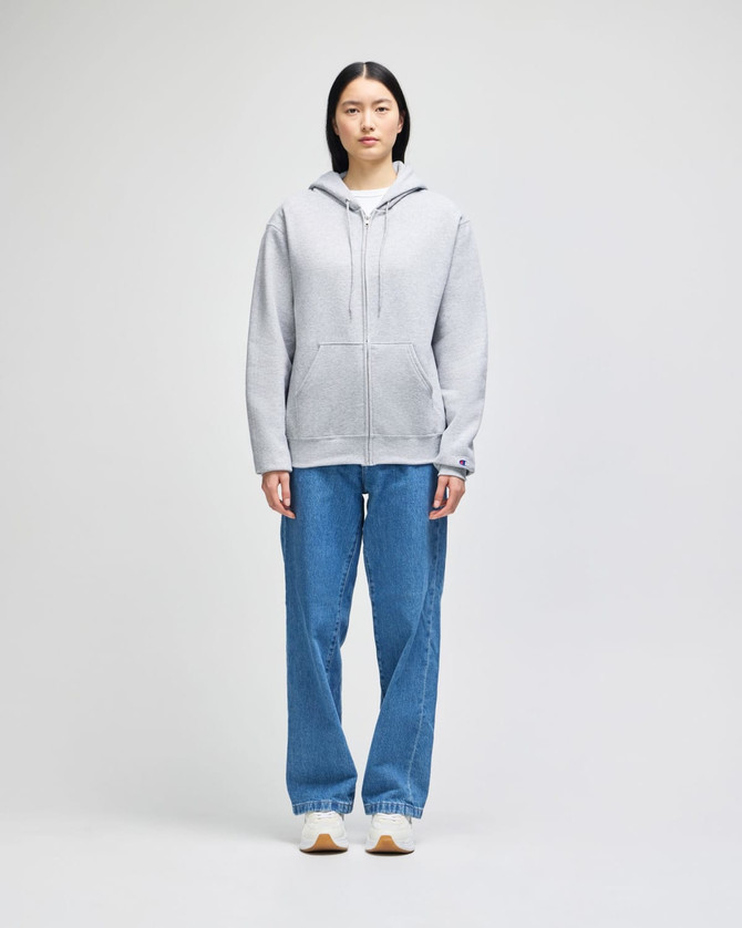 A grey full-zip hooded sweatshirt displayed on a model, paired with blue jeans. The sweatshirt features a logo.