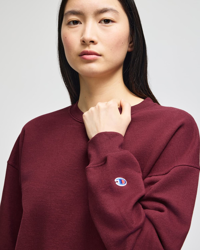 A burgundy women's crewneck sweatshirt featuring a logo on the sleeve. The model is standing and looking forward.