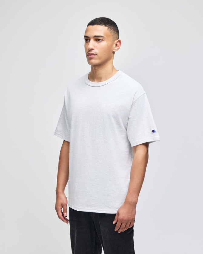 A white Champion adult heritage jersey t-shirt on a model, featuring a logo on one sleeve.