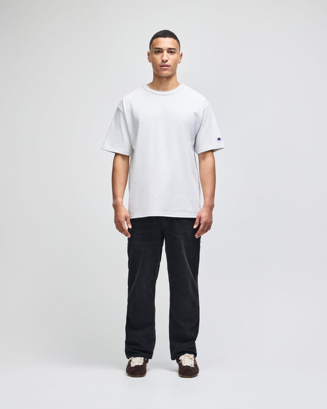 A light grey Champion Adult Heritage Jersey T-Shirt displayed on a model wearing black pants. The shirt has a logo on the sleeve.