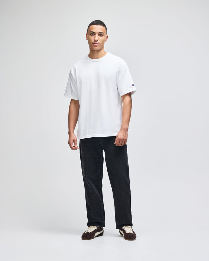 A man wears a white Champion Adult Heritage Jersey T-Shirt with black pants, standing against a plain background.