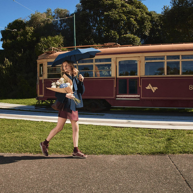 A woman carries a BLUNT Neo Umbrella in navy blue while walking beside a vintage tram in a green outdoor setting.