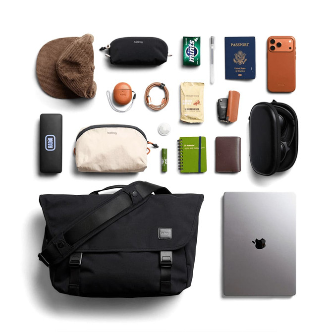 A black messenger bag with various items including sunglasses, a hat, a laptop, and a wallet, arranged on a flat surface.