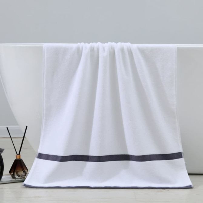 A white bath towel with a dark grey stripe hangs over a bathtub, accompanied by a decorative reed diffuser.