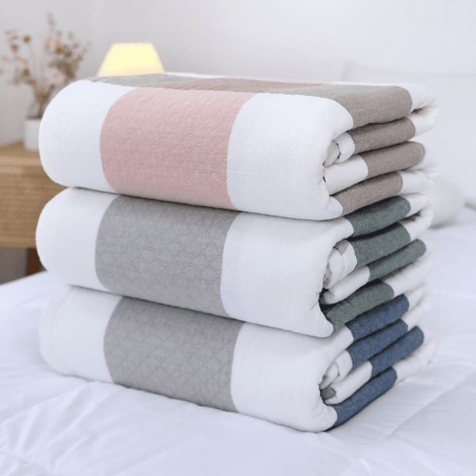 A stack of four blankets in grey, white, pink, and teal, neatly folded on a bed. The image features a logo.