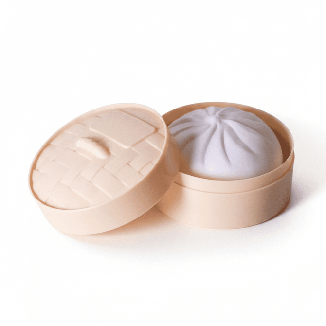 A squishy bun toy in a light peach round container with a lid, resembling a dim sum steamer.