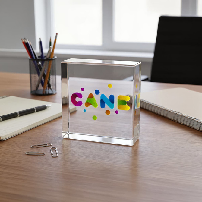 A clear acrylic block with colourful letters and dots, placed on a wooden desk alongside pens, notepads, and paperclips.