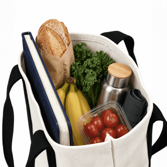 A mini canvas tote in cream filled with a loaf of bread, vegetables, a drink bottle, bananas, and a notebook.