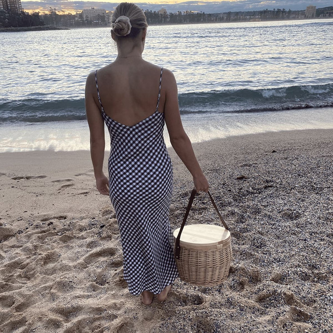 A wicker cooler basket with a wooden lid, carried by a person in a striped dress, on a beach at sunset.