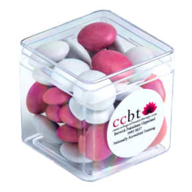 A transparent cube filled with pink and white choc beans, featuring a branded sticker on one side.