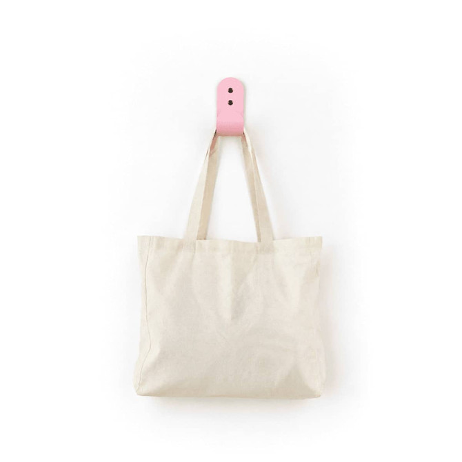 A natural cotton tote bag in a soft cream colour, hanging by its straps on a pink hook.