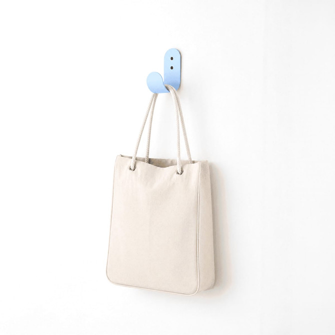 A cream-coloured tote bag hanging on a blue hook against a white wall. The bag has a simple design and a logo.