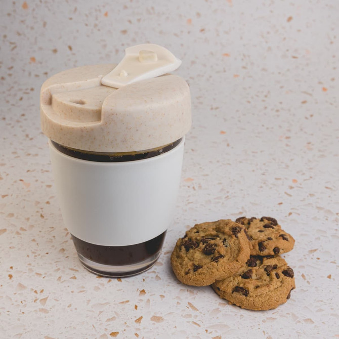 A reusable coffee cup with a beige lid and white body, accompanied by chocolate chip cookies on a speckled surface.