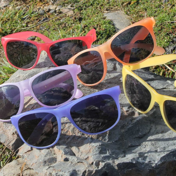 A collection of six sunglasses in pink, orange, purple, and yellow, resting on a rocky surface outdoors.