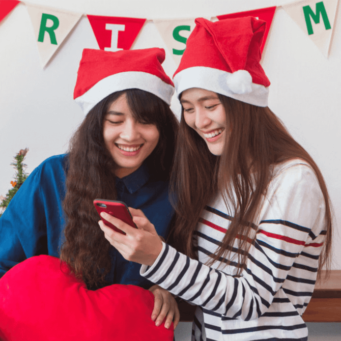 Two women wearing red Christmas Santa hats, smiling and looking at a phone. One holds a red heart cushion.