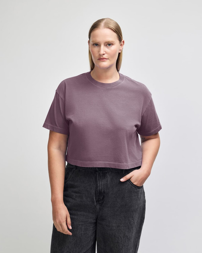 A women's cropped boxy T-shirt in a muted purple shade, featuring a logo. The model is styled with jeans.
