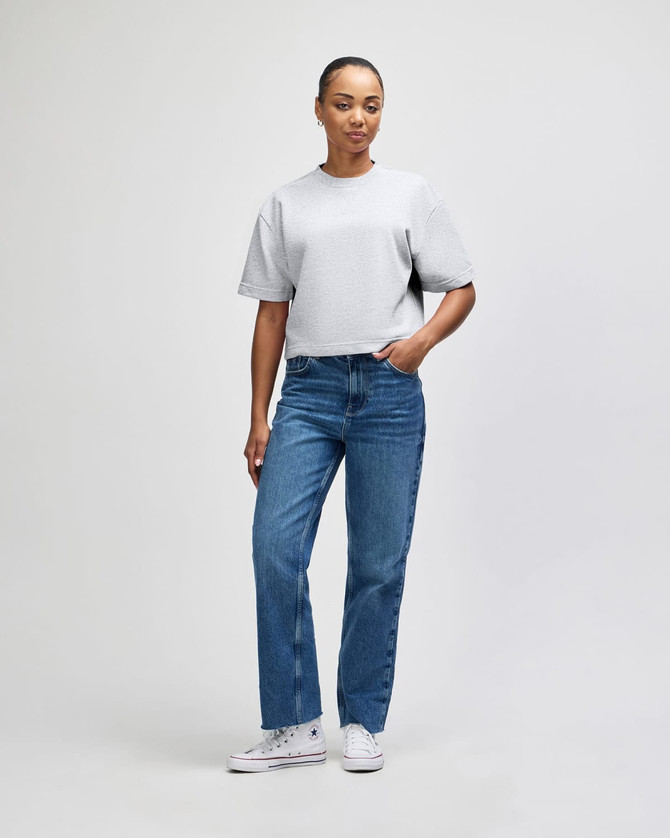 A woman stands in a grey, mid-length boxy top paired with light blue wide-leg jeans, wearing white shoes.