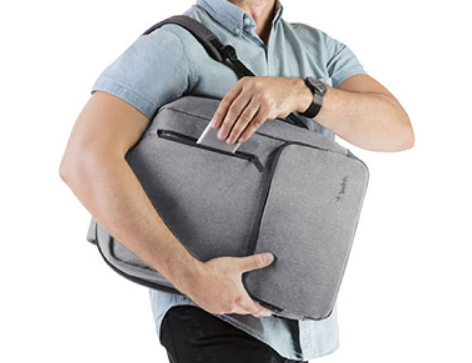 A grey backpack is being carried by a person, with a visible pocket and a logo. The person is wearing a light blue shirt.