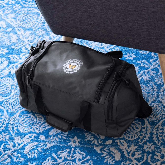 A black duffel bag made from recycled material, featuring a branding logo, resting on a blue patterned rug.
