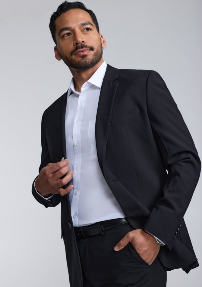 A man in a black suit jacket over a white shirt, standing confidently against a light grey background.