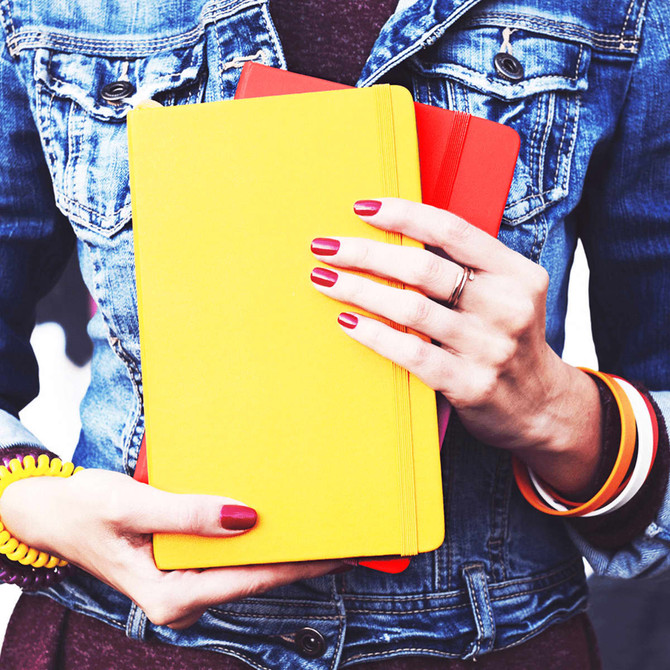 A person holds a yellow notebook along with a red notebook, wearing a denim jacket and colourful bracelets.