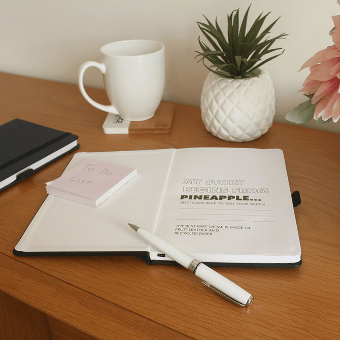A pineapple-themed leather-bound journal with a white pen, a coffee mug, and a small notepad on a wooden surface.