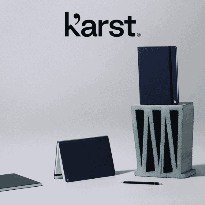 A navy hardcover notebook stands on top of a grey block, accompanied by a smaller notebook and a pen. It has a logo.