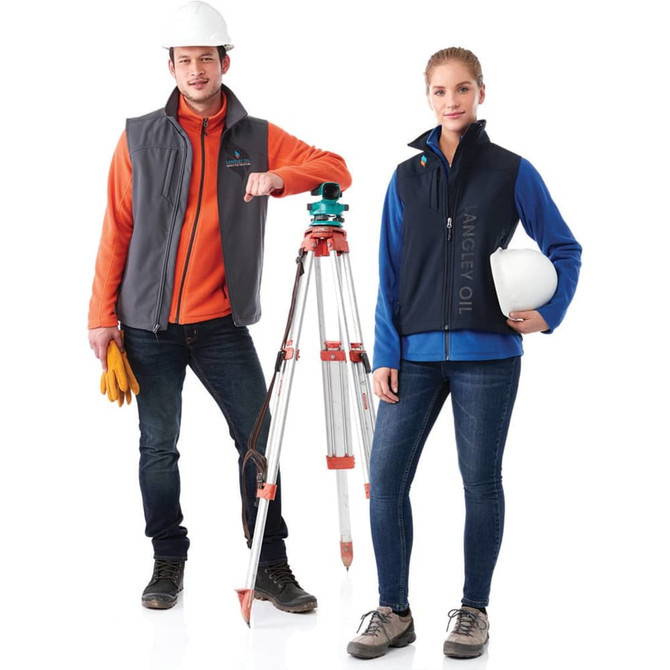 A grey and navy softshell vest is worn by two people, one holding a helmet and the other next to surveying equipment.