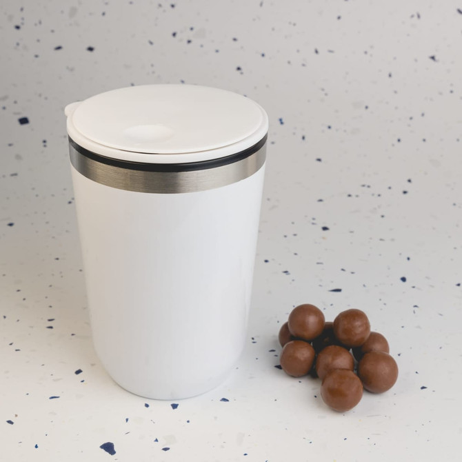 A white reusable coffee cup with a silver band at the top, alongside some brown candies.