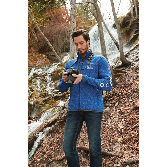 A man in a blue knit jacket stands by a waterfall, holding a camera. The jacket features a logo on one sleeve.