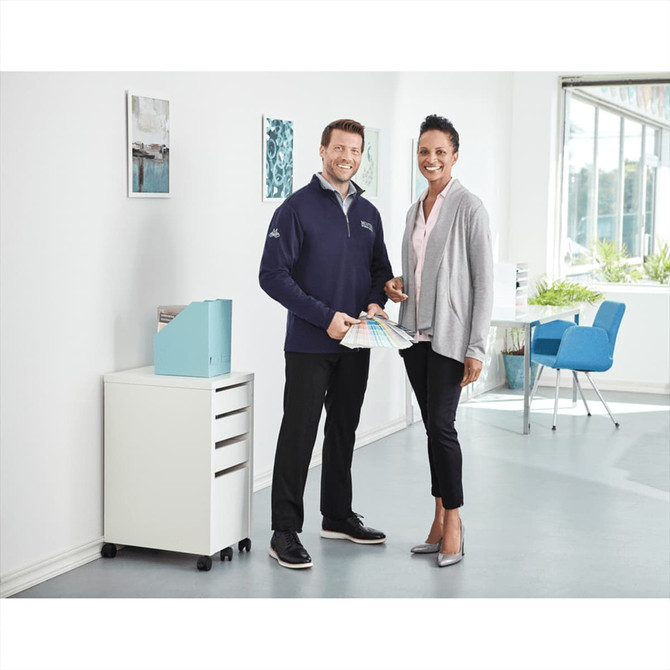 A man in a navy quarter zip jumper and a woman in a grey cardigan stand together, holding fabric swatches in a bright office setting.