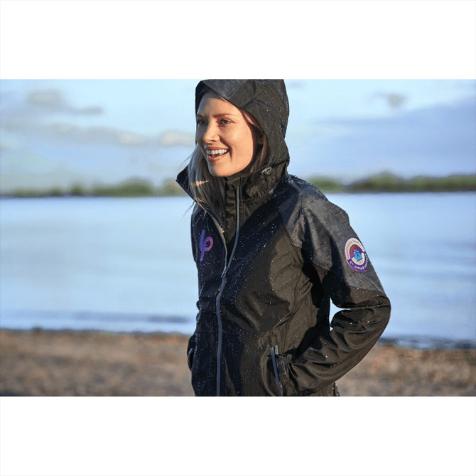 A woman wearing a black softshell jacket with a hood, featuring a logo, standing by a beach with a scenic background.