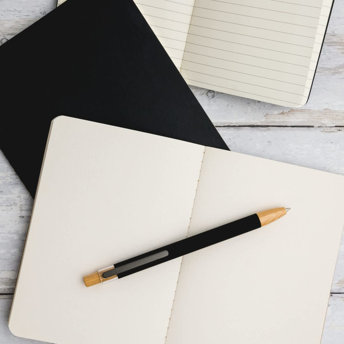A black aluminium pen rests on an open cream notebook, with a black notepad also visible in the background.