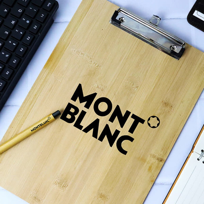 A bamboo clipboard with a dark logo, placed on a surface with a keyboard, pen, and notebook.