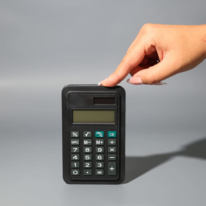 A black calculator with a display and buttons, being held by a hand against a grey background.
